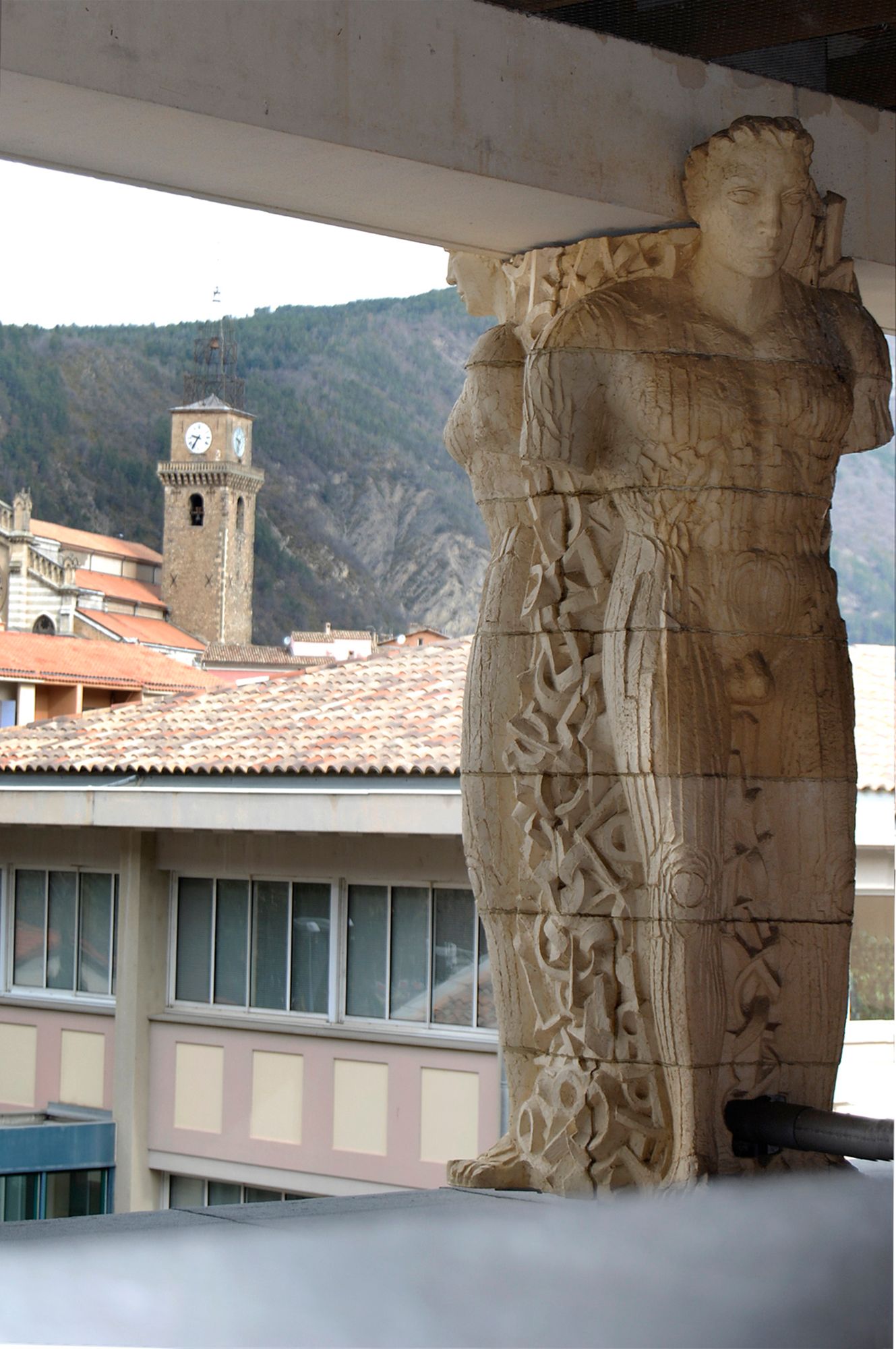 Cariatide du bâtiment des Archives, en arrière plan le clocher de Digne.